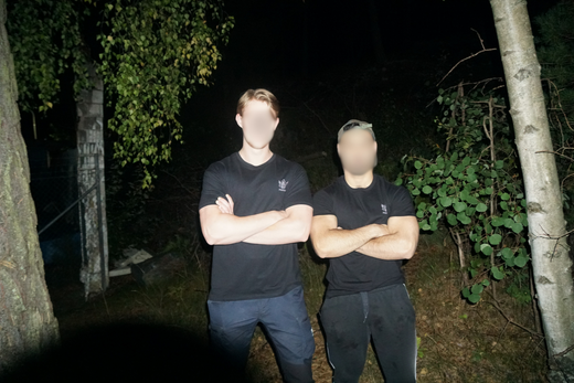 Två personer i svarta träningskläder står med armarna i kors i en mörk skogsmiljö, upplysta av kamerablixt.