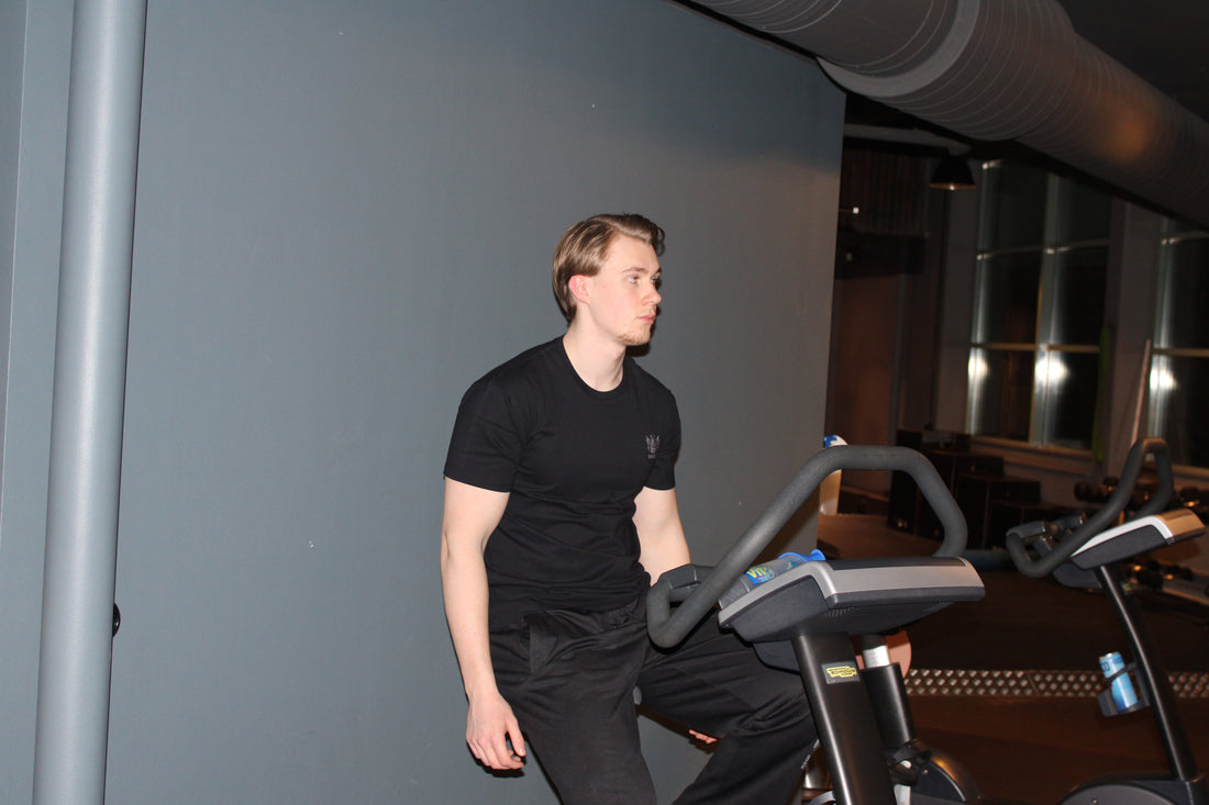 Man i svart träningskläder sitter på en motionscykel på gymmet, med en fokuserad blick och avslappnad hållning. Bakgrunden består av en mörk vägg och stora fönster som reflekterar ljus.