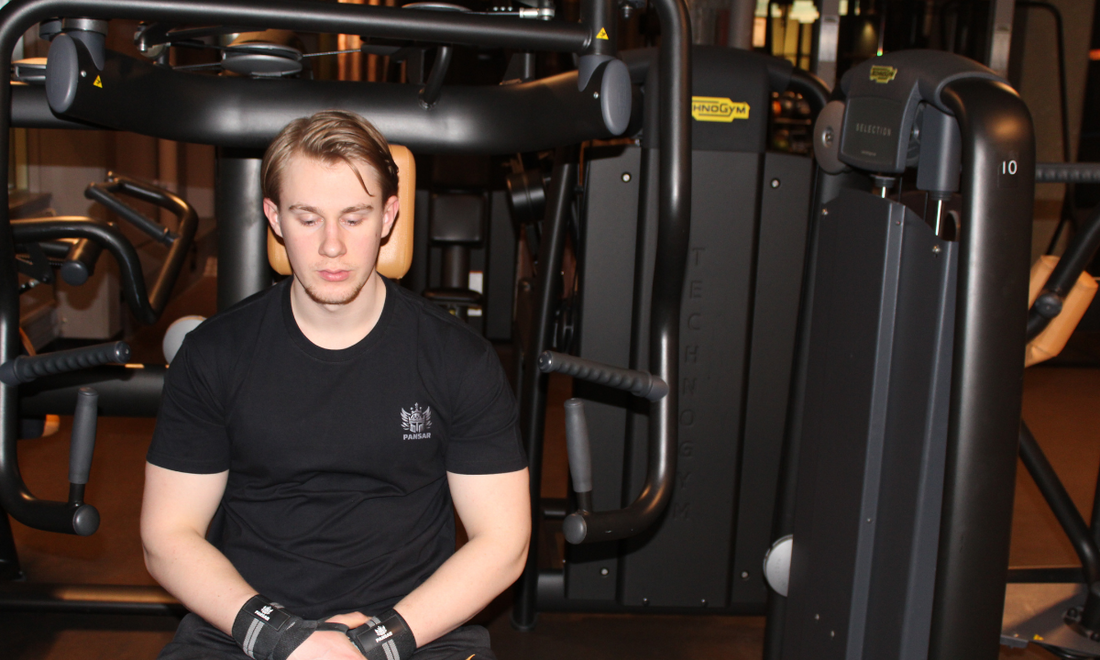 Man i träningskläder från PANSAR sitter fokuserat på gymmet, omgiven av träningsmaskiner – ett ögonblick av mental förberedelse och disciplin.