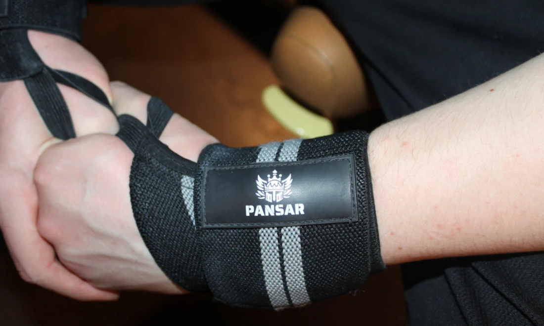 Närbild av en persons handleder iklädda svarta och grå handledslindor med PANSAR-logotypen, redo för tung styrketräning.