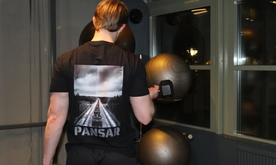 Man med kort, bakåtkammat hår tränar biceps med hantel i gymmiljö, iförd en svart PANSAR-tröja med ett tryck av en militärkolonn på ryggen.