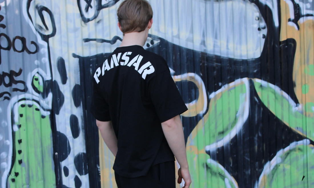 Person bakifrån iklädd svart t-shirt med texten "PANSAR" i vitt, stående framför en färgglad graffitivägg.
