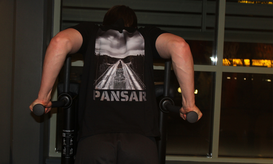 En person tränar triceps-dips på gymmet iförd en svart t-shirt med tryck av en militärkolonn på landsväg och texten "PANSAR" på ryggen.