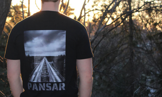 En person står med ryggen mot kameran i en skogsmiljö vid solnedgång. Personen bär en svart t-shirt med ett tryck av ett militärt fordonskonvoj på en landsväg och texten "PANSAR" i blockbokstäver under motivet.