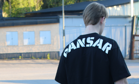 Person i svart t-shirt med texten "PANSAR" på ryggen, fotograferad bakifrån i ett industriområde med mjukt kvällsljus.